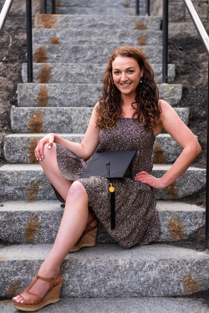 A recent college graduate smiles for the camera during a graduation photoshoot. The background is a beautiful old stone staircase. Logan, UT